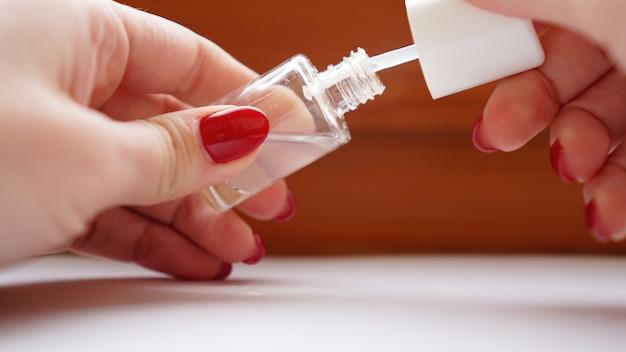 Applying clear nail polish from a small bottle with a red-manicured hand.