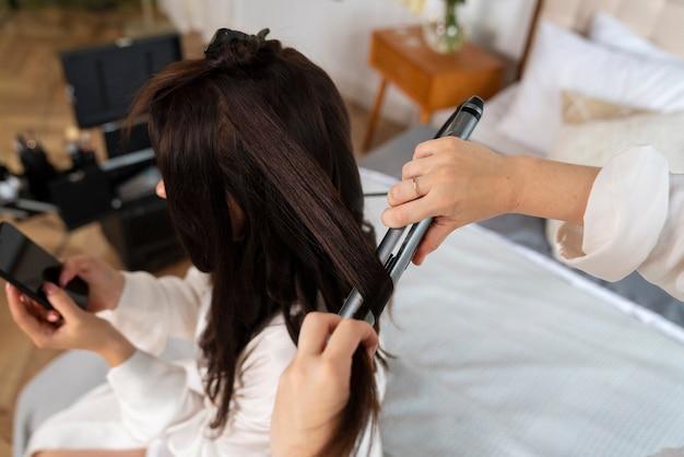 Hairdresser styling a woman's hair with curling iron while client uses smartphone